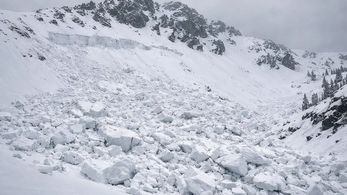 Wide-angle view of a steep snow-covered Sierra Nevada mountain slope showing a large avalanche debris field with fractured slab snow blocks spread across the mountain face below a rocky peak under an overcast winter sky, depicting the aftermath of a major backcountry avalanche near Castle Peak, California.