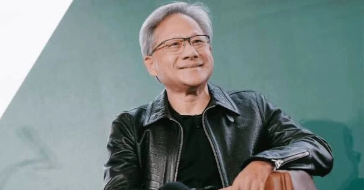 Nvidia CEO Jensen Huang wearing a black leather jacket on stage at a technology conference, presenting new AI hardware to a large audience in a dimly lit auditorium.
