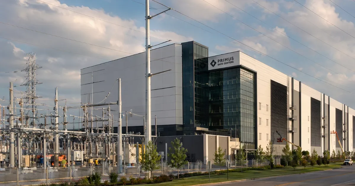 Wide view of a modern data center next to power transmission infrastructure in an industrial setting.