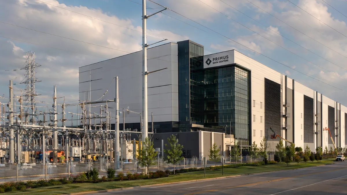 Wide view of a modern data center next to power transmission infrastructure in an industrial setting.