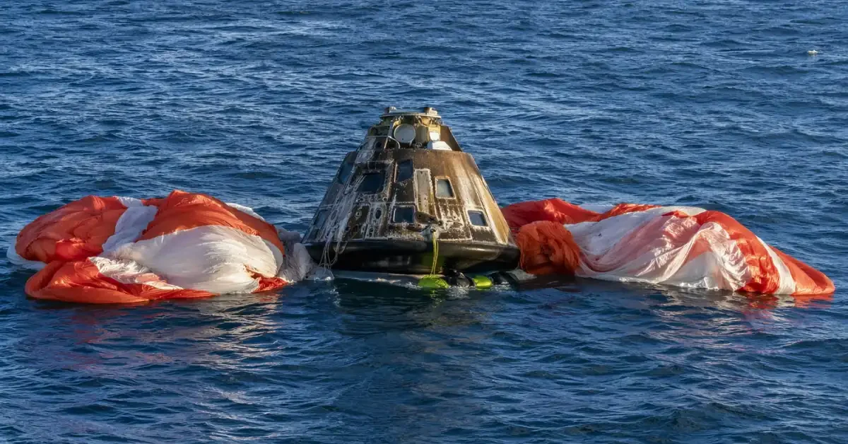 The charred Orion space capsule floating stably in the Pacific Ocean alongside large orange and white parachutes following the historic Artemis II moon mission.
