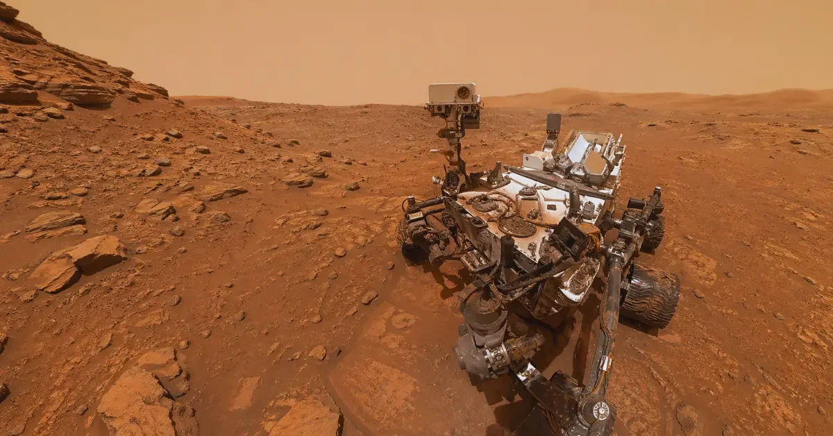 NASA’s Curiosity rover is in a rocky area of Mars with layered red-brown terrain in a wide landscape view.