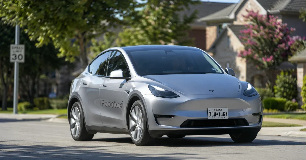 A silver driverless Tesla Model Y driving down a sunlit suburban neighborhood street.